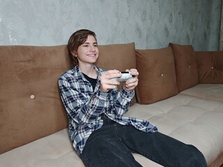 Teenager Playing Video Game on Sofa. Smiling teenage boy holding a game controller while sitting on a sofa. Relaxed indoor gaming moment. © Anastasiya