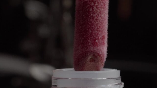 Macro close up of a red textured makeup applicator being inserted into a white cosmetic container, highlighting its soft surface and close contact with the product opening