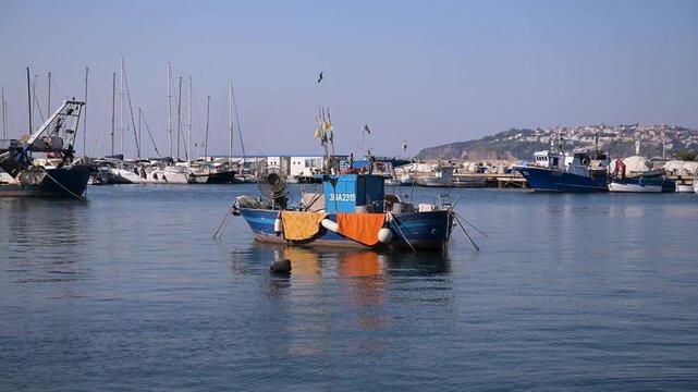 Small blue fishing boat with bright nets floats in a quiet Italian marina with yachts and hillside houses in the background.