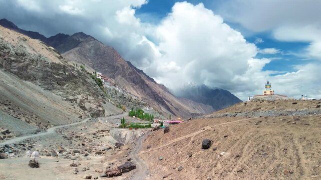 Wide aerial push in shot revealing the giant Maitreya Buddha statue and Diskit Monastery perched on a cliff face under dramatic clouds in Nubra Valley, Ladakh, India.
