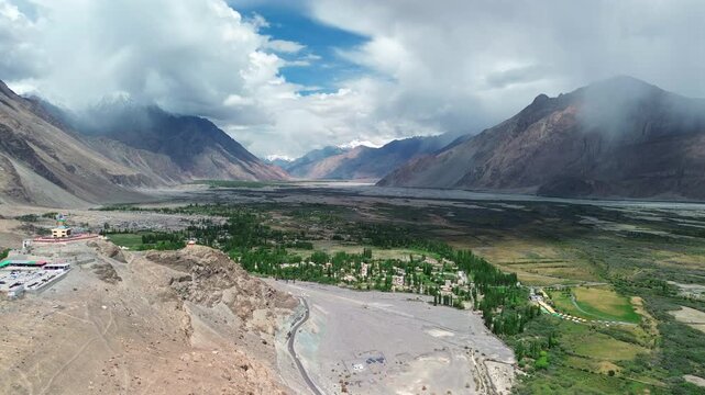 Extreme wide aerial static shot of the expansive Nubra Valley featuring Diskit Monastery, lush green oasis, and dramatic storm clouds over Himalayan peaks in Ladakh, India.
