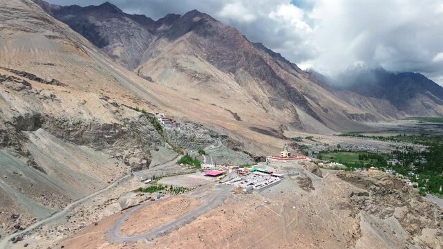 Wide aerial arc shot of Diskit Monastery and the giant statue of Maitreya Buddha surrounded by arid Himalayan mountains in Nubra Valley, Ladakh, India.