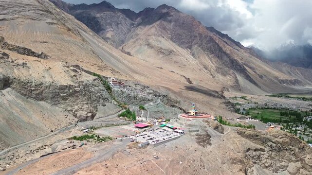 Extreme wide aerial static shot of the Diskit Monastery complex and giant Maitreya Buddha statue nestled against steep rocky mountains in Nubra Valley, Ladakh, India.