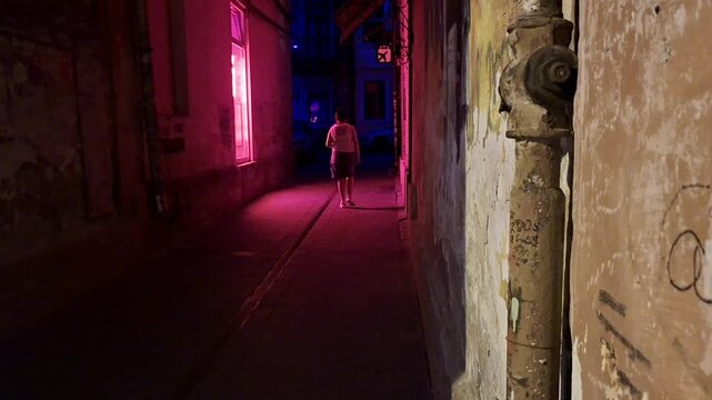 A person walks through a narrow historic alley at night, illuminated by vivid pink and blue neon lights, with weathered walls and atmospheric urban details creating a mysterious mood