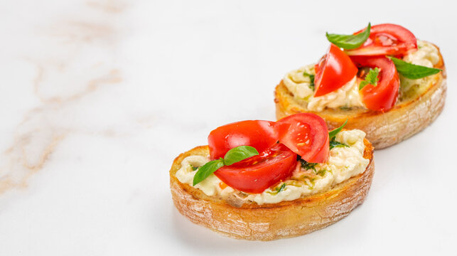 Sliced toasted bread topped with creamy spread, fresh tomato slices, and basil leaves arranged on a marble surface for a gourmet appetizer presentation