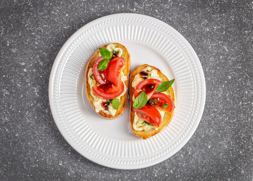 Two slices of toasted bread topped with fresh tomatoes, basil leaves, and creamy cheese served on a white plate against a textured gray background