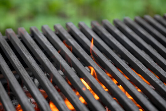 Close-up view of a black metal grill grate with flames and glowing embers beneath, set against a blurred green background in an outdoor cooking environment
