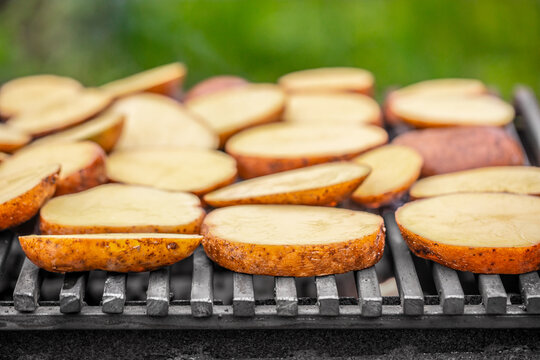 Slices of potatoes grilling on a barbecue grill with a blurred green background, showcasing a summer cooking scene with food preparation outdoors