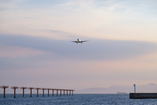 夕暮れの海と進入灯へ向かって着陸態勢に入る旅客機。淡い青からピンクへ移り変わる涼しい空の色が美しく、夏の終わりのような旅情を誘う風景です。旅行やビジネス、航空関連の背景素材やデザインのアクセントとして最適です。コピースペースも広く、多様なレイアウトに活用できます。