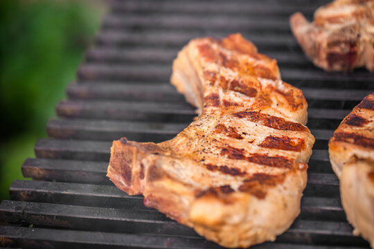 Grilled pork chops with distinct grill marks cooking on a barbecue grill, smoke rising, surrounded by a blurred green outdoor background