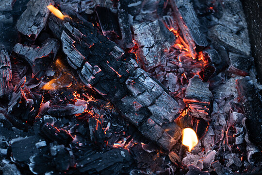 Charred wood logs and glowing embers in a fire pit, showcasing the remnants of a recent fire with ash and glowing orange highlights visible
