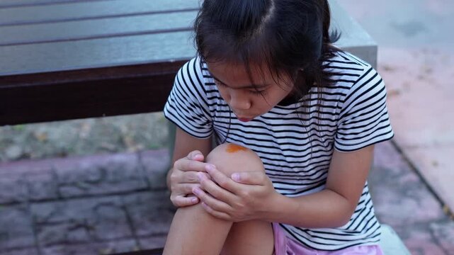 Sad Asian girl sitting and touching injured bleeding knee with painful expression outdoors