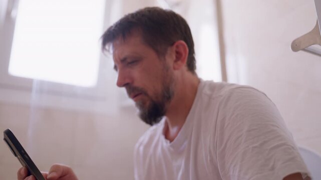 Adult man sitting on toilet and looking at smartphone during a private moment, showing a satisfied facial expression while engaging in masturbation and self-stimulation.