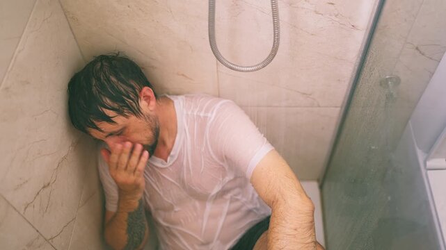 Sad man  sitting alone and fully clothed in the corner of a shower under running water, struggling with depression, anxiety, grief, and despair