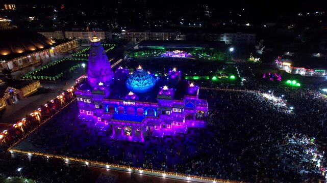 Aerial drone view of the beautifully illuminated Prem Mandir in Vrindavan, India, during Janmashtami celebrations at night. The magnificent temple glows with colorful lights that continuously change.