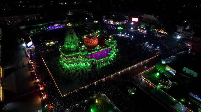 Aerial drone view of the beautifully illuminated Prem Mandir in Vrindavan, India, during Janmashtami celebrations at night. The magnificent temple glows with colorful lights that continuously change.