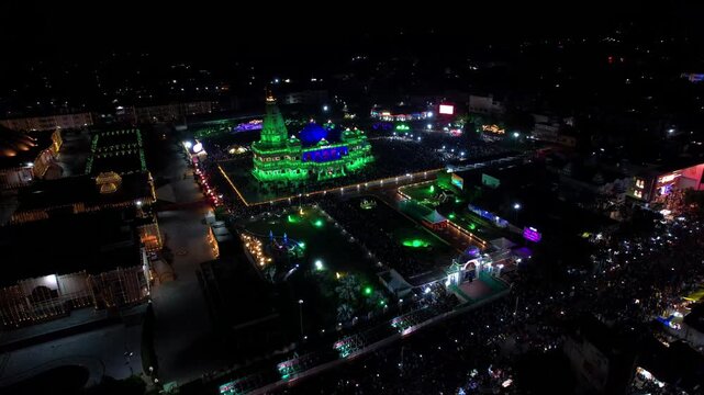 Aerial drone view of the beautifully illuminated Prem Mandir in Vrindavan, India, during Janmashtami celebrations at night. The magnificent temple glows with colorful lights that continuously change.