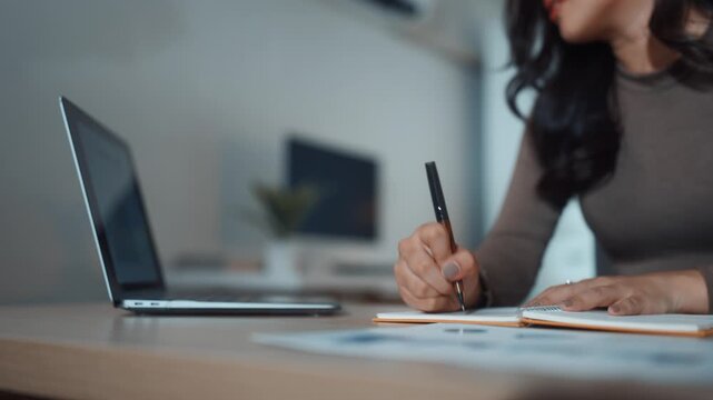 Focused businesswoman writing notes in a notebook while working on a laptop at her desk in a modern office at night