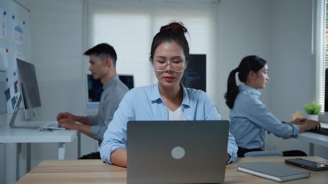 Young Asian female programmer thinking and solving a coding problem on her laptop computer with colleagues in the office