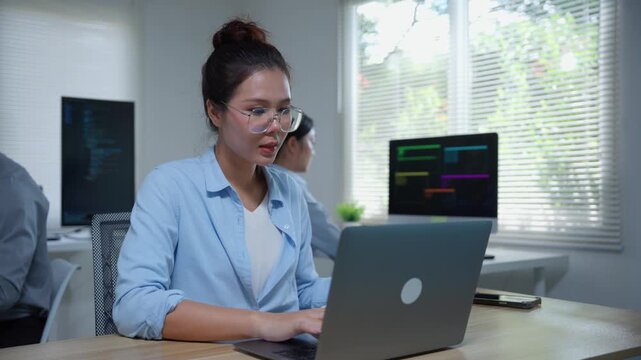 Asian female programmer developing software code on a laptop computer with her team in an office