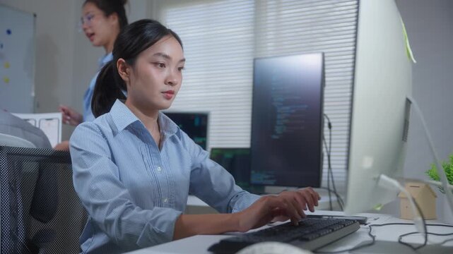 Asian female software developer coding on a desktop computer while working with a team of colleagues in a busy office setting