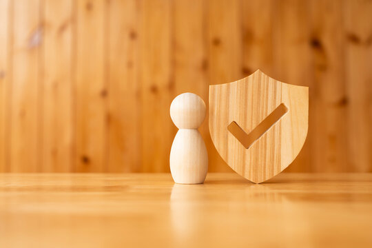 Life insurance and personal protection concept with wooden doll figure standing next to wood shield with check mark icon, representing approved guarantee, safety assurance, and secure coverage.