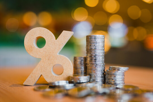 Interest rate and compound growth concept with wooden percentage sign beside stack of money coins on warm bokeh background, representing bank deposit dividend, financial savings, and tax return.