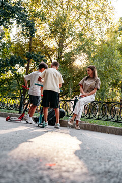 Children playing with scooters in a park while an adult watches nearby in the afternoon sunlight