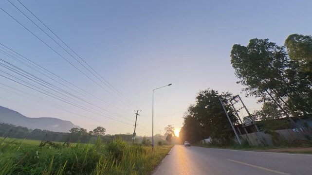 Slow camera pan across a scenic rural road at sunset, revealing green fields, distant mountains, trees and warm golden light in a peaceful countryside landscape.