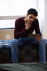 African American man sitting on ottoman by blinds in maroon sweater touching chin holding tablet © WavebreakMediaMicro
