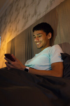 African American man reclining in bedroom on bed, smiling at smartphone while showing smartwatch