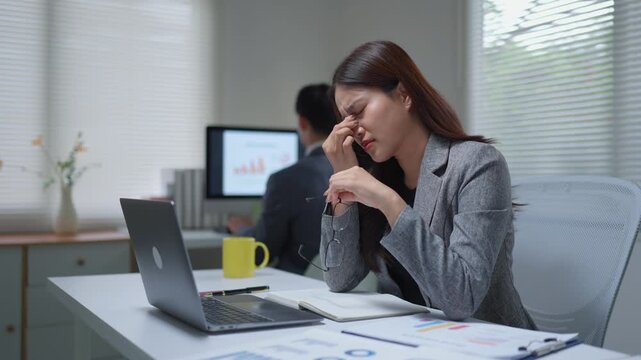Stressed businesswoman with headache and eye strain working on laptop in modern office, feeling overworked and tired