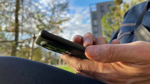 Close up of male hands scrolling smartphone screen in green park. Mature man slides fingers through online social media feed lines outdoors. Casual digital network communication concept