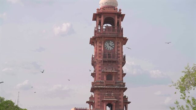 Medium low angle slow motion static shot of the famous red sandstone Ghanta Ghar Clock Tower with birds flying around the landmark in Jodhpur, Rajasthan, India.