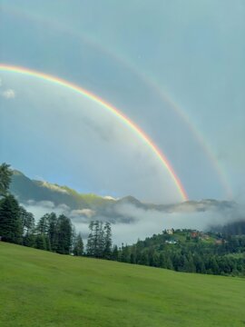 Bright double rainbow arching over mist covered green mountains and rural village valley after rain.