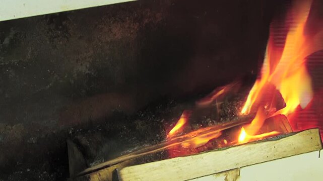 The footage shows burning wood in metal combustion chamber. The view includes orange yellow fire around dark wooden logs and metal ventilation slots within metal combustion chamber.