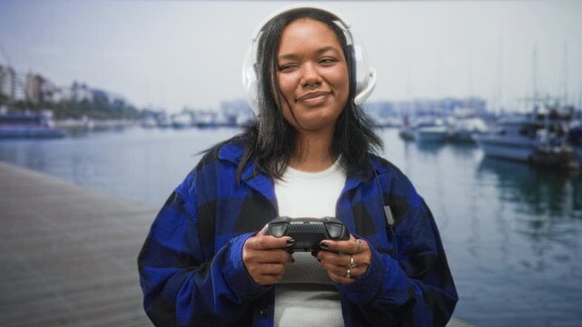 Young woman holding game controller, wearing headset, thumbs on buttons and smiling on a street by a marina dock; joyful relaxed play.