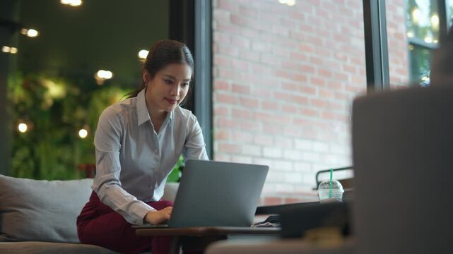 Young Asian woman sitting on a comfortable sofa, working on her laptop. The freelancer is typing and browsing online in a cafe