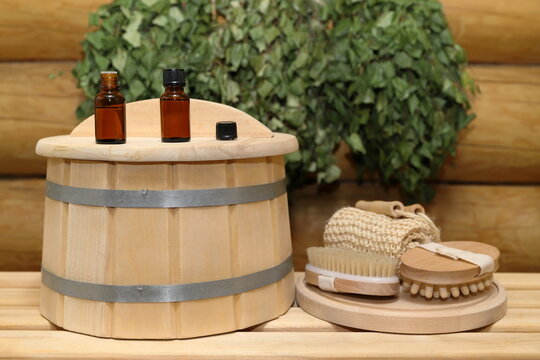 On a plank bench, on top of a wooden barrel, next to set of bath supplies on wooden round tray, there are two bottles of healing essential oil, against the background of log wall with a birch brooms. 