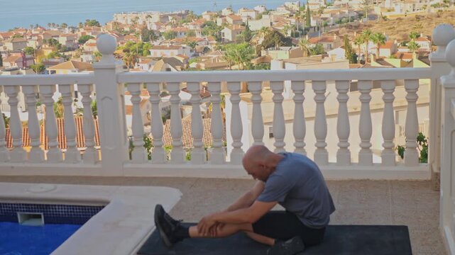 Side view of a middle aged man stretching his legs and back on a fitness mat next to a swimming pool on a balcony terrace with sea views.