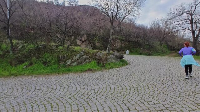 A man and a woman in athletic wear jogging together along a cobblestone road winding up a green hill park in Plovdiv.