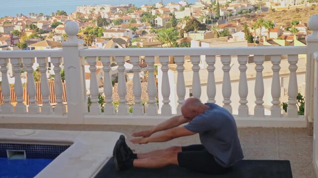 Side view of a middle aged man stretching his hamstrings while seated on a yoga mat on a pool terrace with coastal background views.