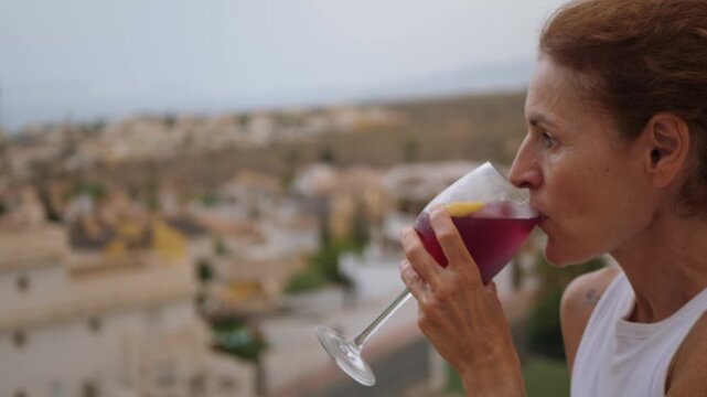 Close up shot of a middle aged woman taking a sip of red sangria cocktail from a wine glass on a balcony terrace with blurred background.