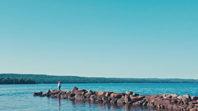 A woman walking along a break wall of rocks out to a massive body of water surrounded by a bay of distant forests and trees in this daring nature shot on a sunny summer day.