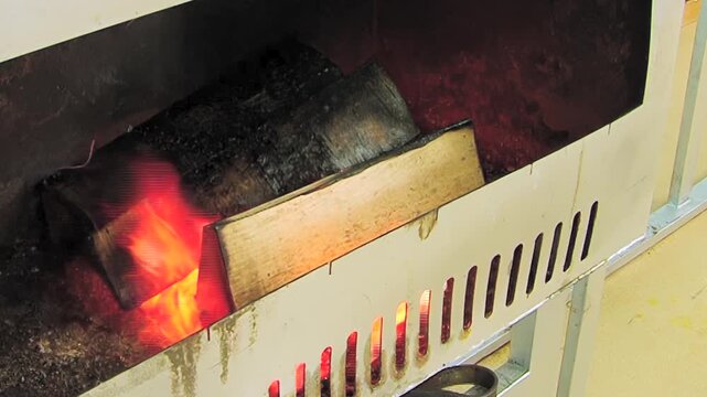 The footage shows burning wood in vented metal chamber. In the frame, orange yellow flames move around dark wooden logs beside metal ventilation slots within vented metal chamber.