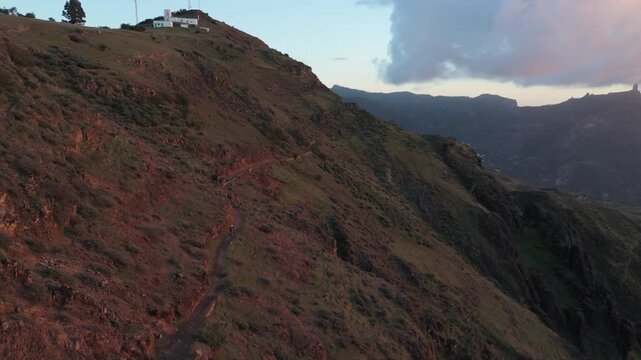 A drone push in shot revealing the rugged mountains and trails of Gran Canaria at sunset. A lone hiker makes their way along a dirt path towards the Cruz de Tejeda hamlet.