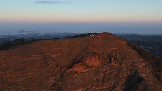 A aerial drone shot pans right over a rugged mountain peak on Gran Canaria at sunset. The volcanic landscape is bathed in warm orange light, with communication towers and distant city lights.