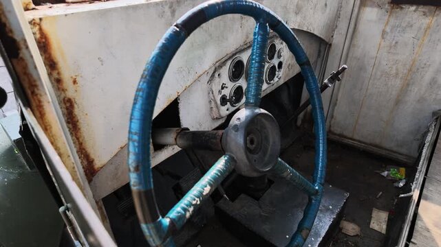 A weathered blue steering wheel with rusted metal components sits centered in a dilapidated vehicle cabin as the camera slowly pans right to reveal the deteriorated dashboard and side panel.