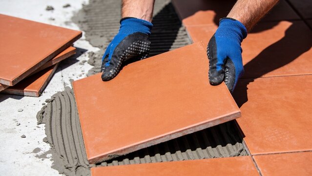 Professional tiler positioning terracotta ceramic floor tile over freshly combed gray adhesive mortar using blue protective gloves precise placement alignment spacing joints flooring installation bath
