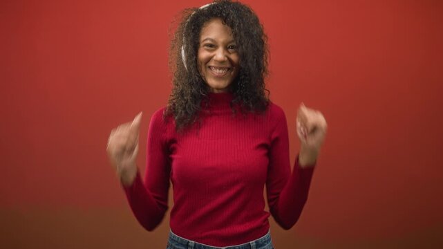 Teenage woman with curly hair wearing headphones smiles, claps and pumps hands while dancing in a red studio set; joy celebration music.